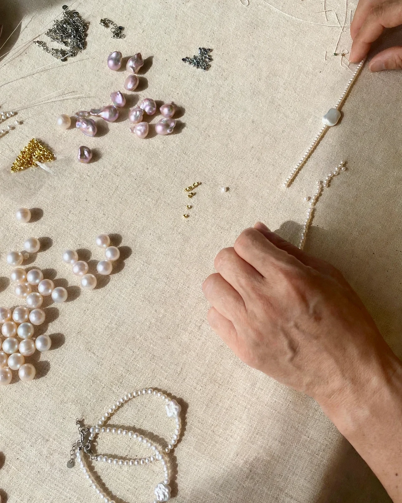 Loose pearls, chain and findings on a linen workbench, a hand stringing a strand in sunlight